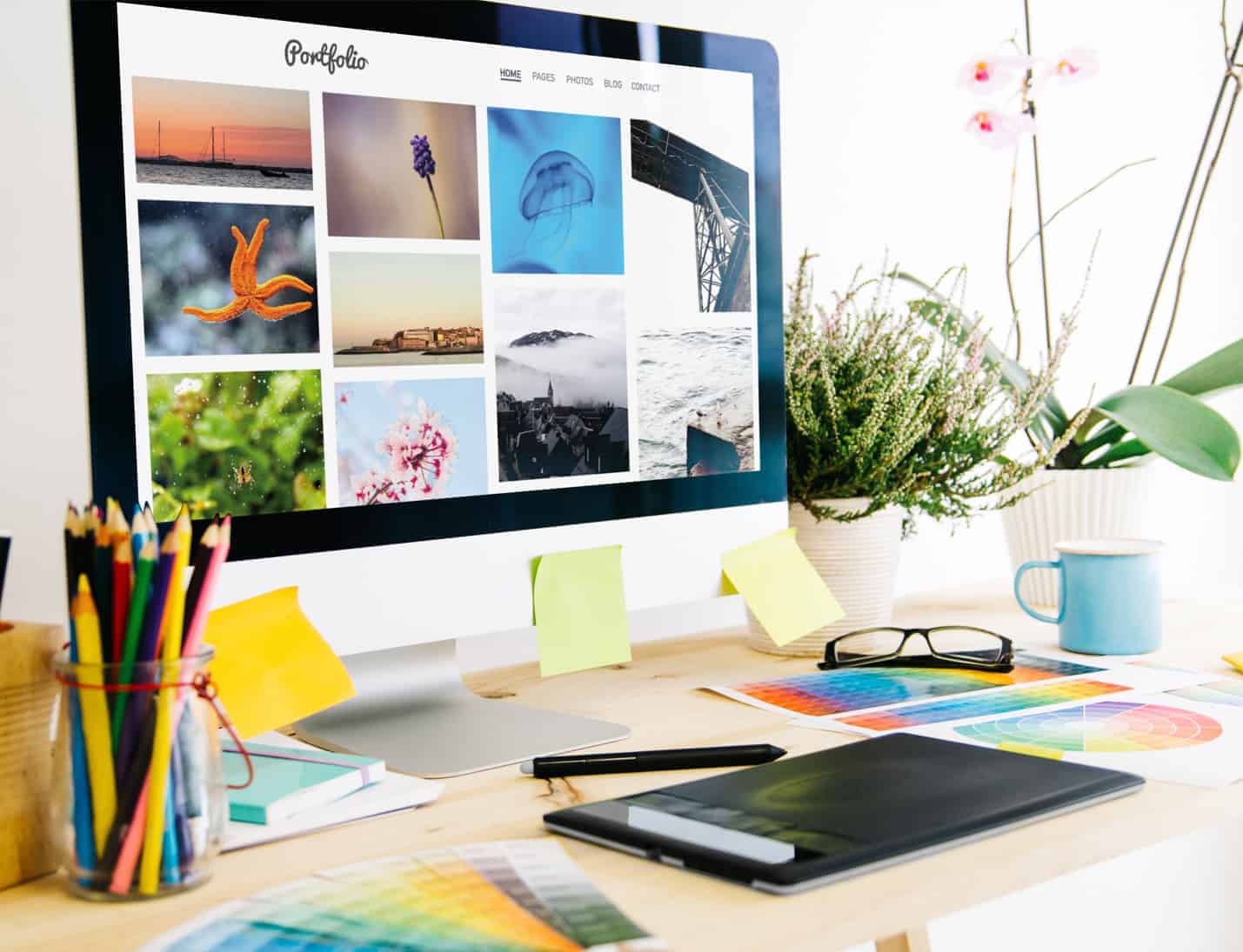 about EWD (Esthetig Web Design) about us page: web design that works for you. Image shows a desk with a computer screen showing a portfolio on it. The view is from 45 degree angle to the desk, on the desk are various stationery items, colour swatches, plants and glasses.