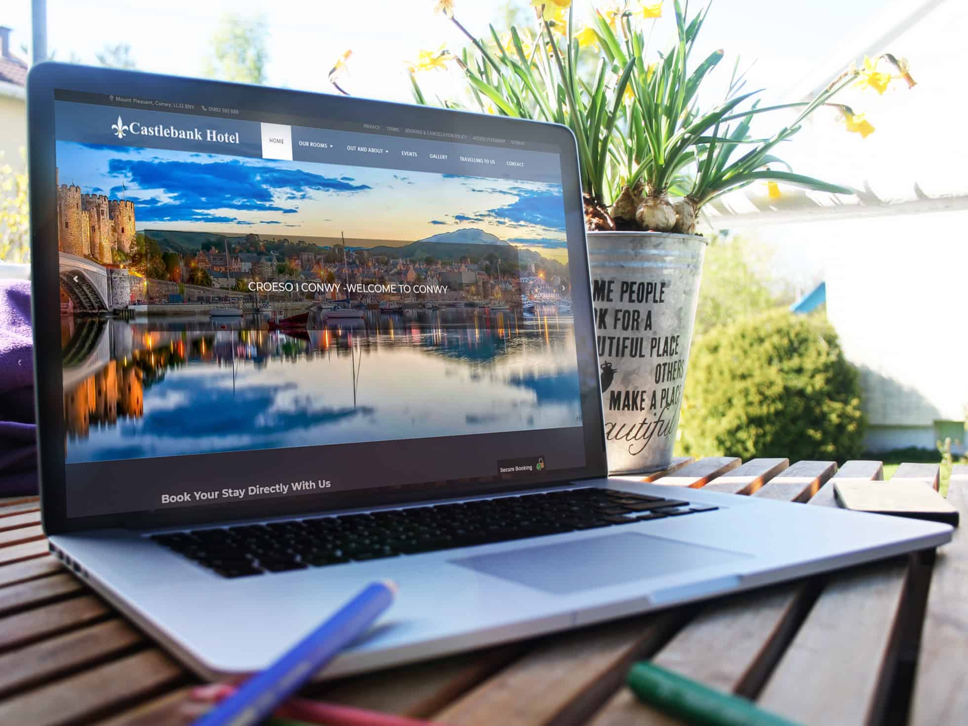 EWD (Esthetig Web Design) Portfolio: Castlebank Hotel, website shown on a Macbook Pro sitting on a slatted table in a garden, to the right of the Macbook is a plant in a silver bucket with wording on it.