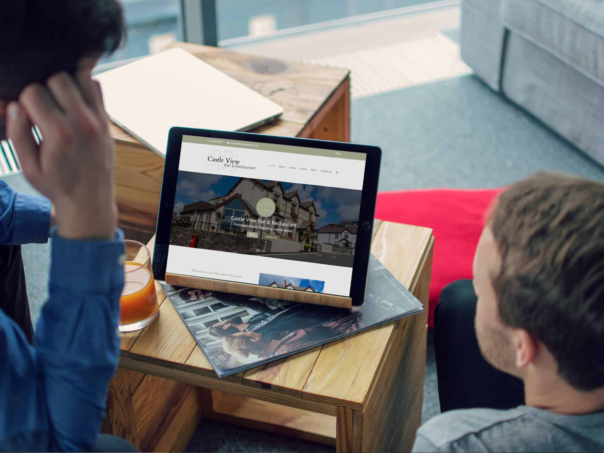 EWD (Esthetig Web Design) Portfolio: Castle View Pub, website shown on an iPad Pro that is sitting on a table on a stand being viewed by two men.