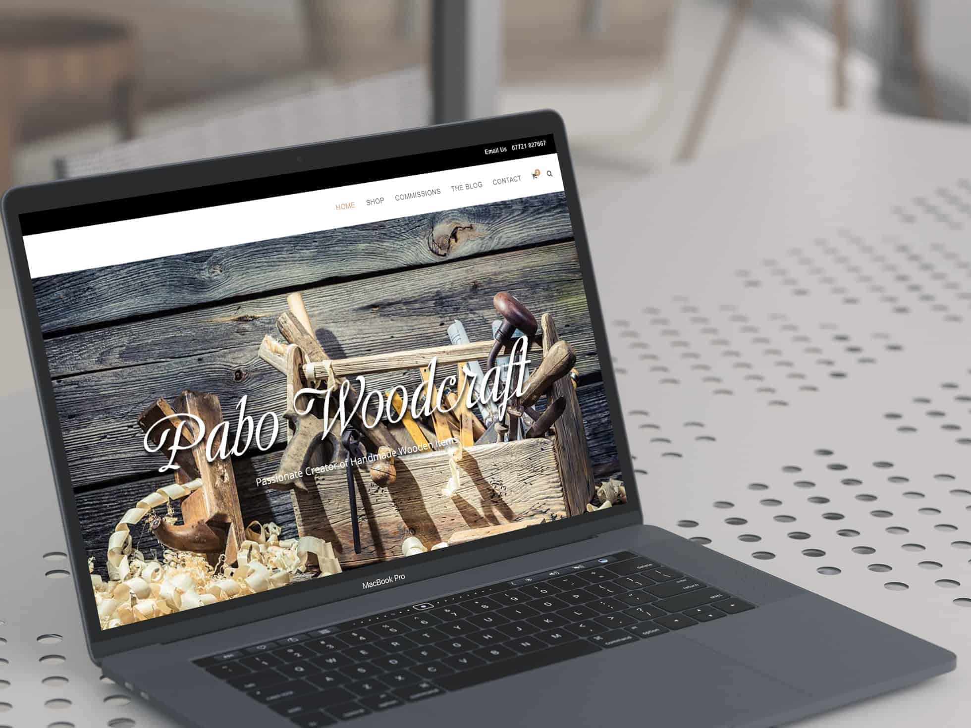 EWD (Esthetig Web Design) Portfolio: Pabo Woodcraft, website shown on an Macbook Air placed on a white table that has circular cutouts.