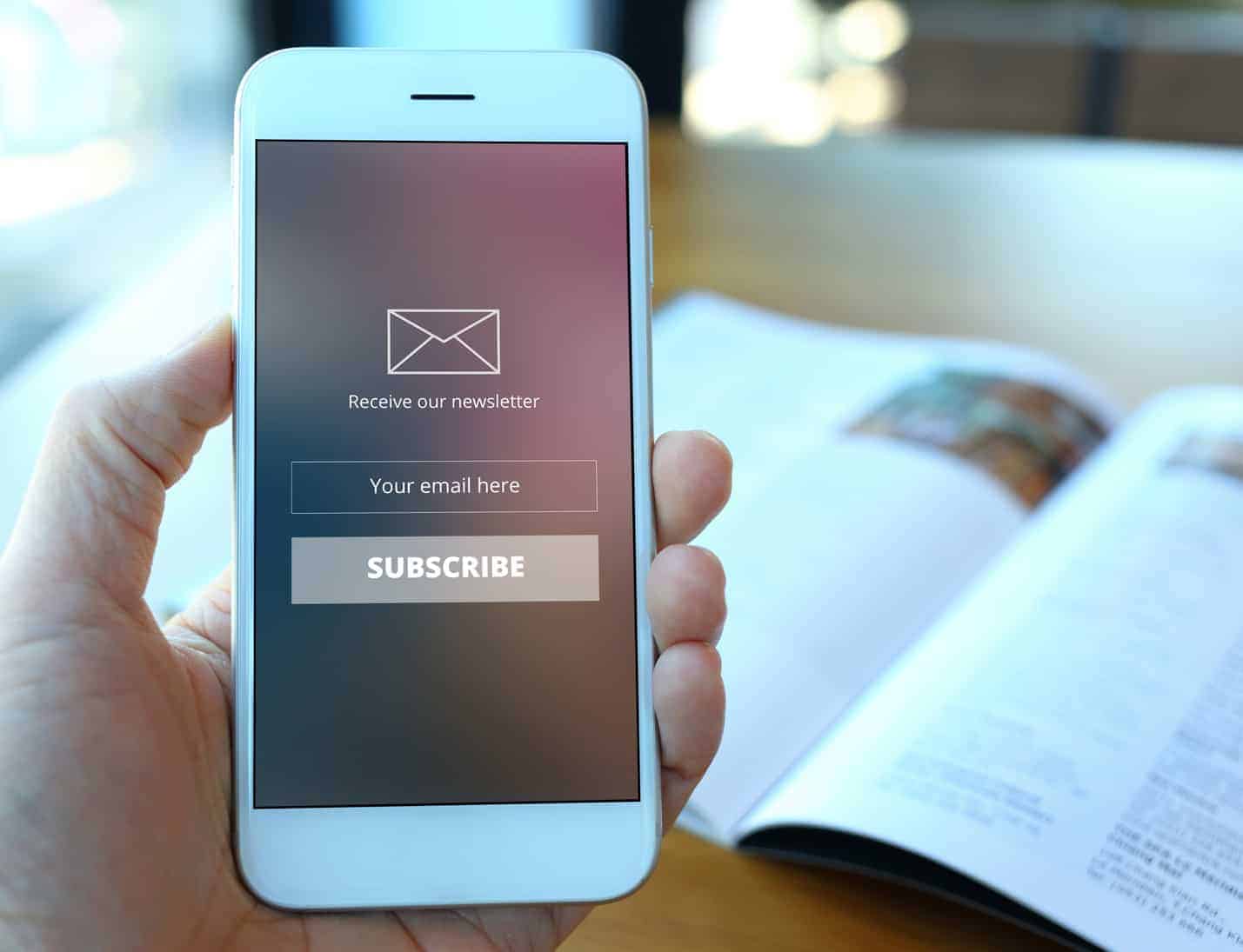 Photo of a person holding a mobile phone above a table. On the mobile screen is a newsletter subscription form. The phone itself is held over a table with an open magazine on it.