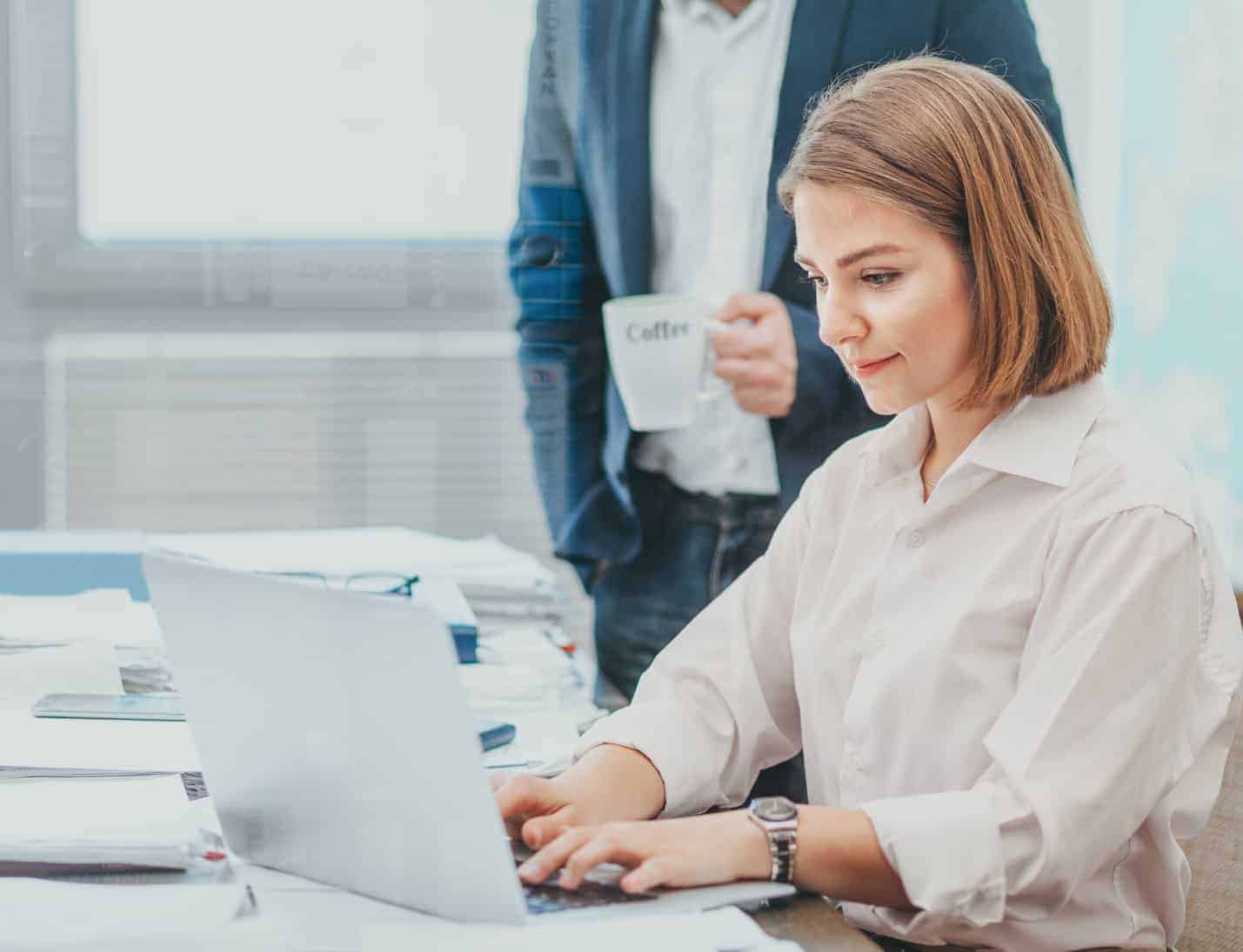GDPR and email marketing. Woman using a laptop on a table, with stacks of files to her right. Standing over her right should is a man holding a mug and watching what she is doing. The woman seems unaware he is looking. The image is quite bright, the woman is wearing a white shirt and the man (head not showing) is wearing jeans a jacket and white shirt.