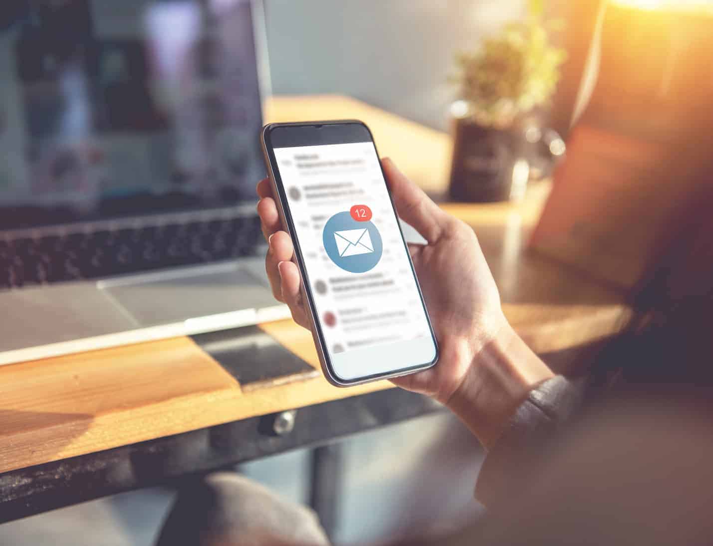 EWD (Esthetig Web Design) email marketing. Photo of a person (blurred out) sitting in front of a laptop on a desk and holding a mobile phone. On the phone is an open email box with a blue circle icon in the middle of the screen with a white envelope and a small red circle with a count of the emails. To the right of the laptop is a coffee cup and diffused light coming through the window.