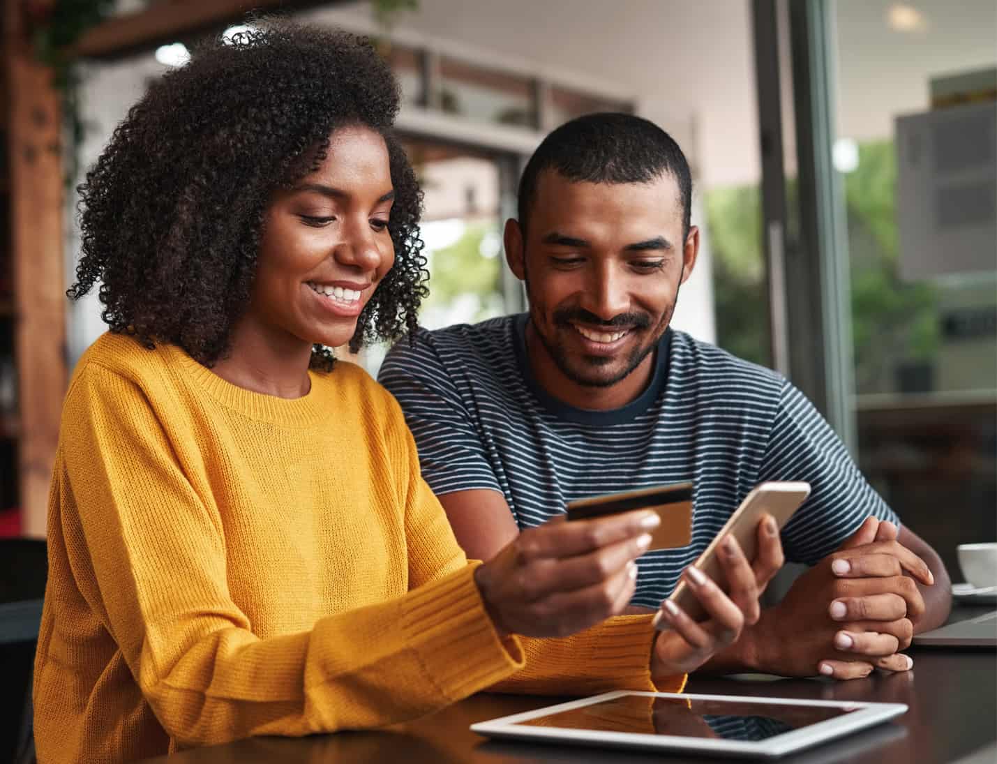 blog post: responsive web design. Couple using mobile devices to make a purchase, woman holding mobile phone with a card in one hand ready to buy with her partner looking on.